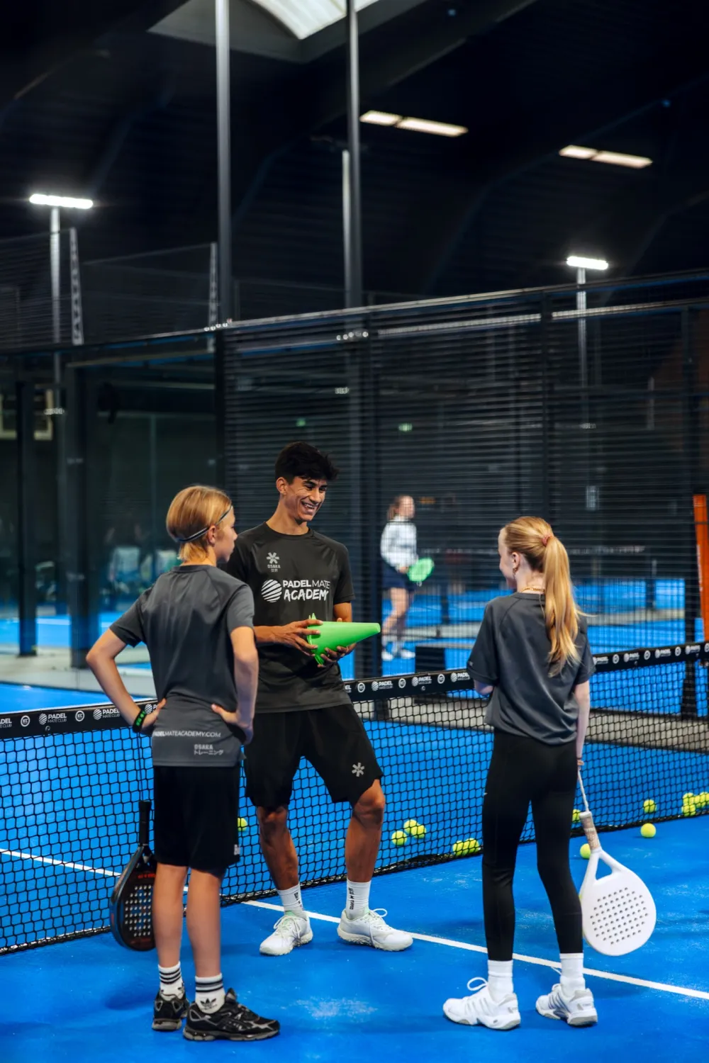 Trainer van Padel Mate Academy lacht met twee jonge leerlingen aan het net